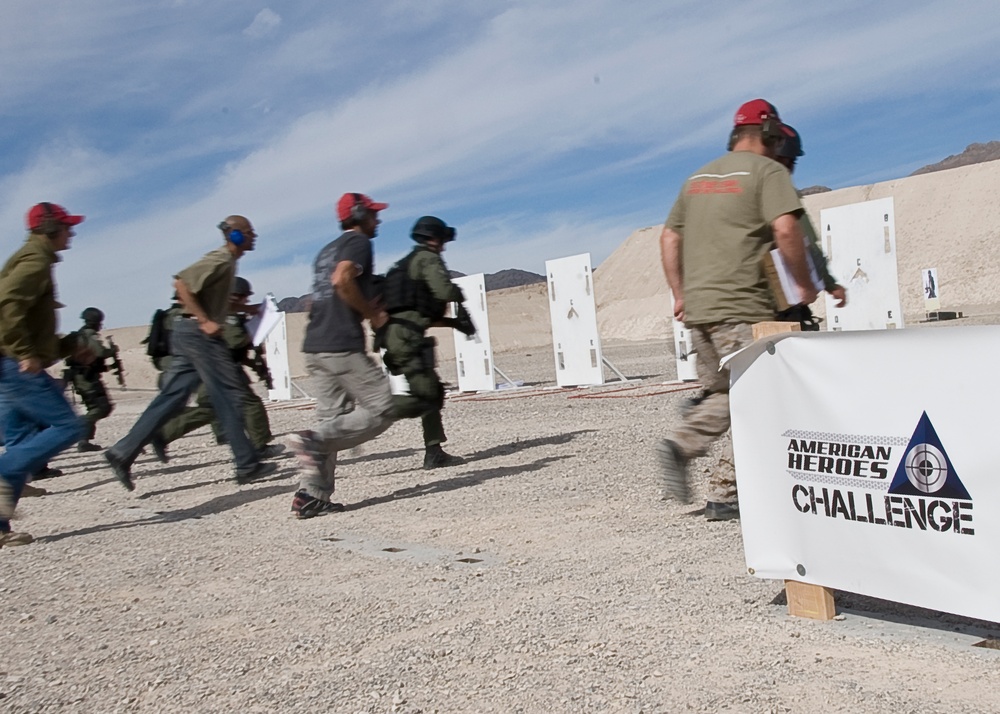 A group of participants are running at an outdoor shooting range during the 'American Heroes Challenge' competition. The event appears to take place in a desert environment with white target stands visible in the background. Participants are wearing red caps, hearing protection, and tactical gear.