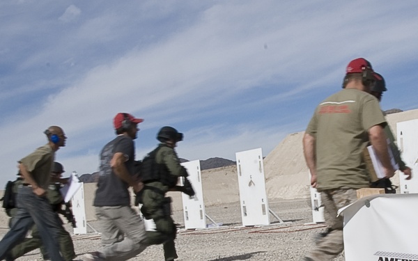 A group of participants are running at an outdoor shooting range during the 'American Heroes Challenge' competition. The event appears to take place in a desert environment with white target stands visible in the background. Participants are wearing red caps, hearing protection, and tactical gear.
