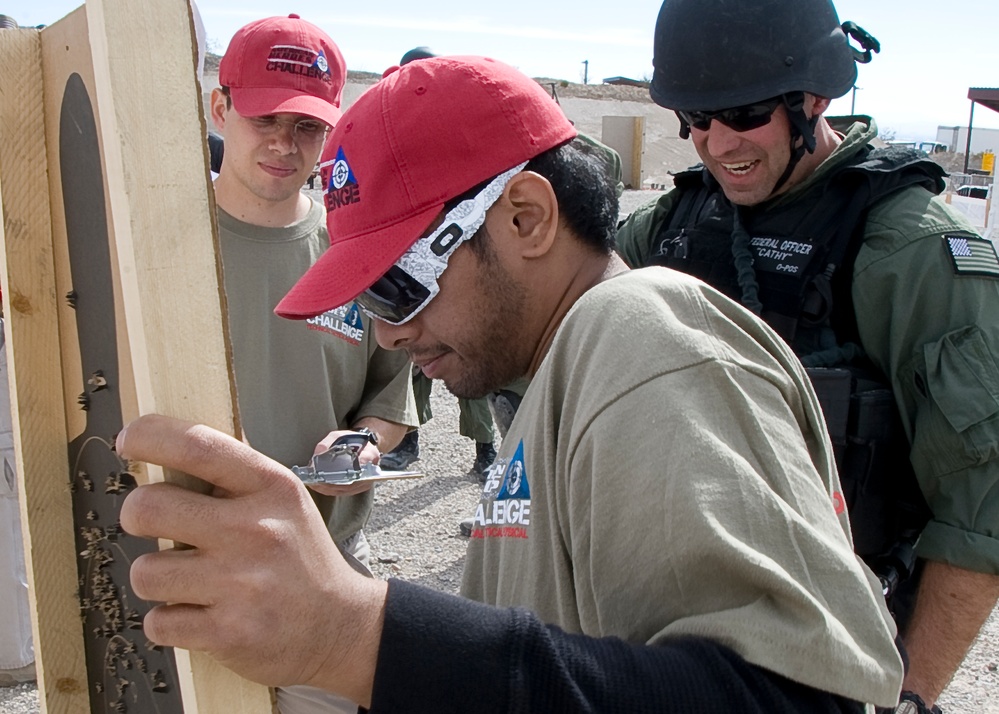 A group of people are examining what appears to be a shooting target at an outdoor range. The central figure wearing a red cap and sunglasses is closely inspecting bullet holes in a dark metal or cardboard target mounted on a wooden frame. A law enforcement or security officer in tactical gear and helmet is observing alongside civilians wearing matching 'Challenge' branded shirts.