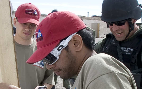 A group of people are examining what appears to be a shooting target at an outdoor range. The central figure wearing a red cap and sunglasses is closely inspecting bullet holes in a dark metal or cardboard target mounted on a wooden frame. A law enforcement or security officer in tactical gear and helmet is observing alongside civilians wearing matching 'Challenge' branded shirts.
