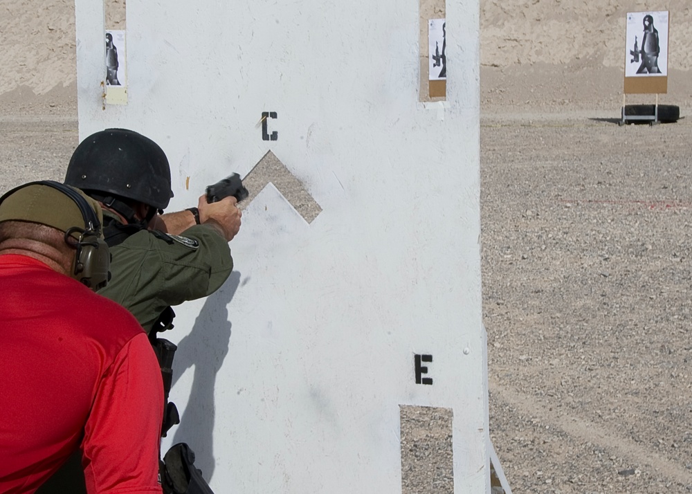 A firearms training exercise is taking place at an outdoor shooting range in an arid environment. A shooter in tactical gear and helmet is firing a pistol around a white barricade wall, while a second person in a red shirt observes from behind. Multiple paper silhouette targets are positioned downrange at varying distances.