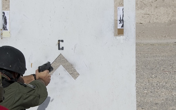 A firearms training exercise is taking place at an outdoor shooting range in an arid environment. A shooter in tactical gear and helmet is firing a pistol around a white barricade wall, while a second person in a red shirt observes from behind. Multiple paper silhouette targets are positioned downrange at varying distances.