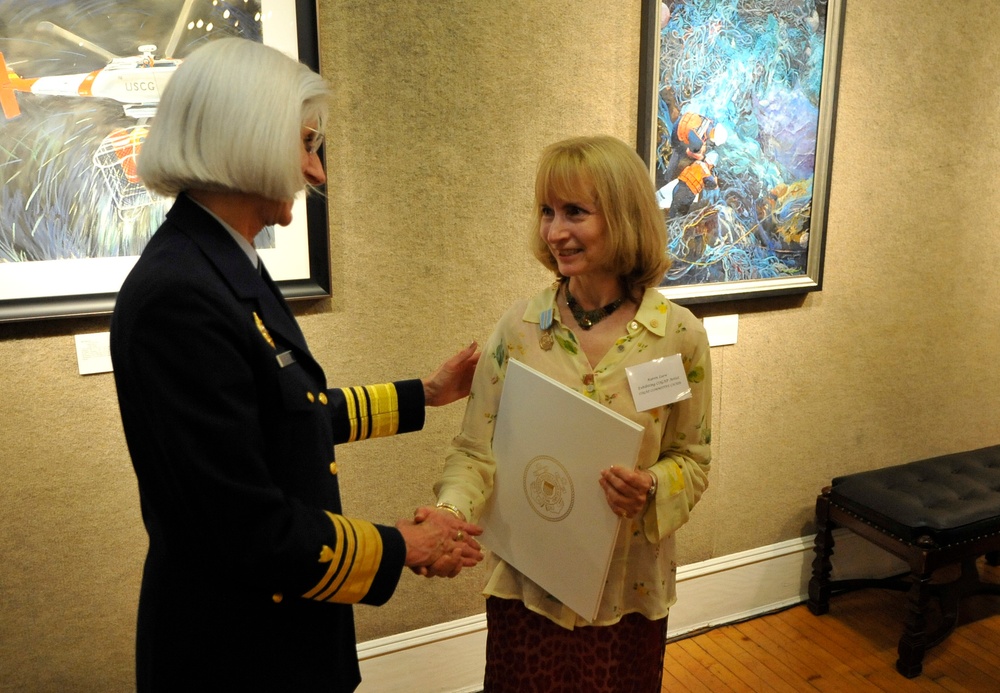 A military officer in a dark navy uniform with gold sleeve stripes is shaking hands with a civilian woman in a floral blouse. The civilian woman is holding a white folder or certificate bearing an official seal. The setting appears to be an art gallery or formal reception room with framed paintings on the walls.