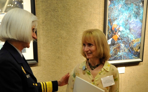 A military officer in a dark navy uniform with gold sleeve stripes is shaking hands with a civilian woman in a floral blouse. The civilian woman is holding a white folder or certificate bearing an official seal. The setting appears to be an art gallery or formal reception room with framed paintings on the walls.