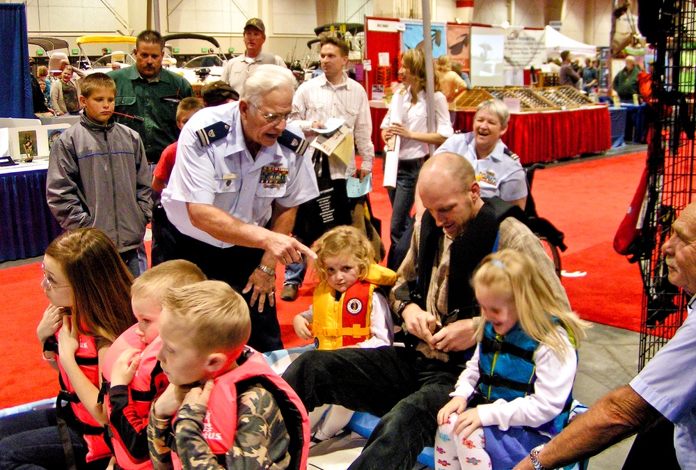 An indoor expo or boat show where a uniformed military officer (wearing medals and insignia) is demonstrating life jackets to children and families. Multiple children are being fitted with orange, red, and blue life vests, suggesting a water safety demonstration booth. The event appears to be a large convention center with multiple vendor displays and red carpet flooring visible in the background.