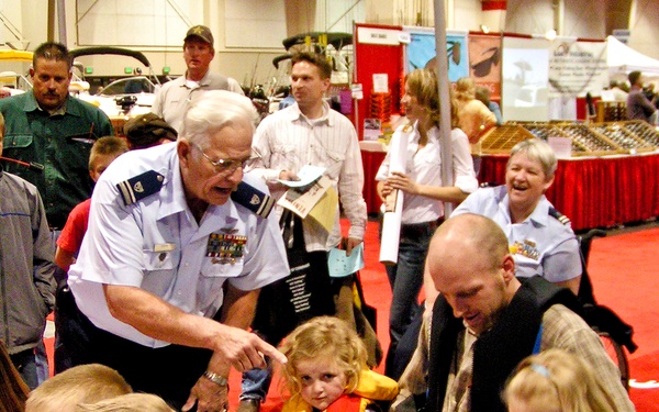 An indoor expo or boat show where a uniformed military officer (wearing medals and insignia) is demonstrating life jackets to children and families. Multiple children are being fitted with orange, red, and blue life vests, suggesting a water safety demonstration booth. The event appears to be a large convention center with multiple vendor displays and red carpet flooring visible in the background.