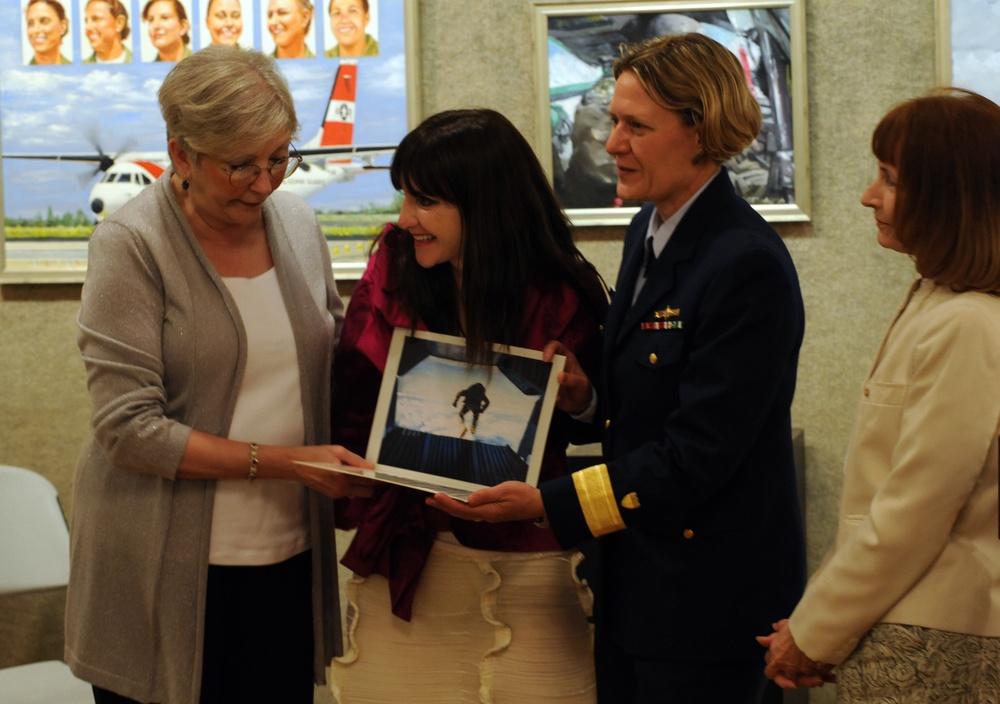 A formal presentation ceremony is taking place indoors, where a uniformed military or coast guard officer is presenting a framed photograph to two civilian women. The officer wears a dark navy uniform with gold sleeve stripes and service ribbons, suggesting a senior rank. The setting appears to be an official building with military-themed artwork and portraits displayed on the walls in the background.