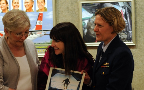 A formal presentation ceremony is taking place indoors, where a uniformed military or coast guard officer is presenting a framed photograph to two civilian women. The officer wears a dark navy uniform with gold sleeve stripes and service ribbons, suggesting a senior rank. The setting appears to be an official building with military-themed artwork and portraits displayed on the walls in the background.