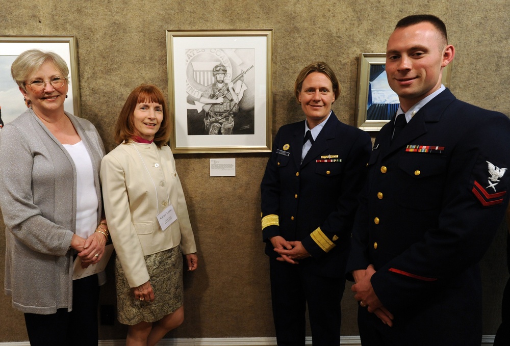 Four individuals pose together in front of framed artwork displayed on a wall, likely at a formal reception or gallery event. Two of the individuals are wearing dark navy dress uniforms with gold buttons and service ribbons, consistent with U.S. Coast Guard attire. The other two are civilian women dressed in business-casual attire.