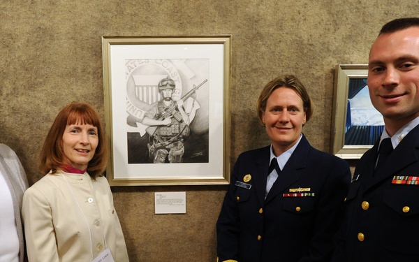 Four individuals pose together in front of framed artwork displayed on a wall, likely at a formal reception or gallery event. Two of the individuals are wearing dark navy dress uniforms with gold buttons and service ribbons, consistent with U.S. Coast Guard attire. The other two are civilian women dressed in business-casual attire.