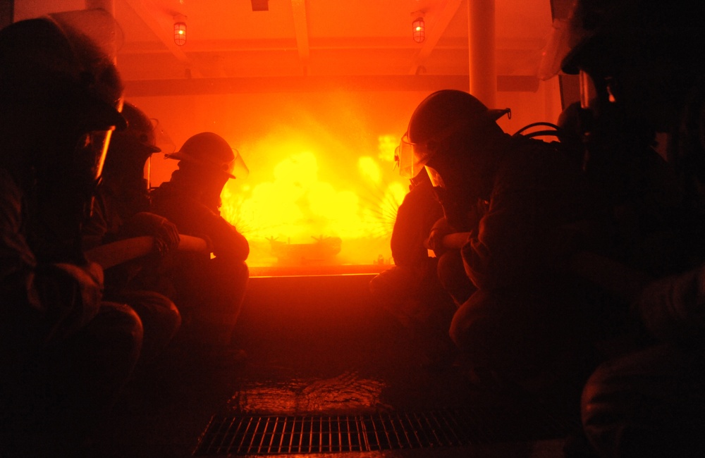 The image shows multiple individuals wearing helmets and breathing apparatus crouched low in a darkened corridor or compartment, facing an intense fire with bright yellow-orange flames visible ahead of them. The scene appears to be a firefighting training exercise or an active firefighting response, likely aboard a ship or in an industrial facility. The environment is filled with smoke and the space is illuminated almost entirely by the fire's glow.