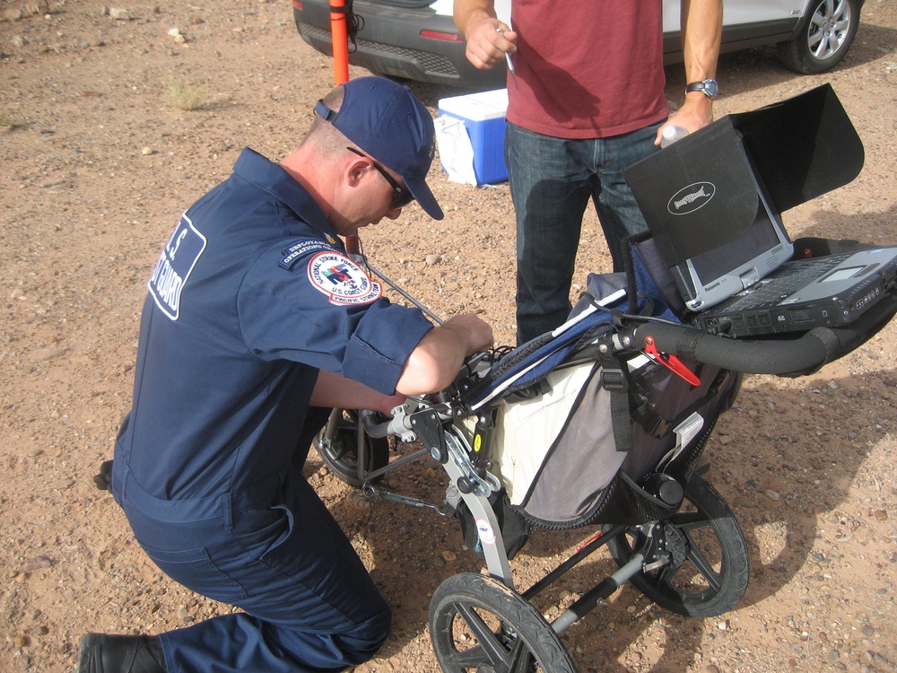 A U.S. Coast Guard member in navy blue uniform is kneeling and working on a wheeled ground-penetrating radar (GPR) cart equipped with a ruggedized Panasonic Toughbook laptop. A second person in civilian clothing (red shirt, jeans) stands nearby observing. The setting appears to be an arid, desert-like outdoor environment with gravel ground and a vehicle visible in the background.