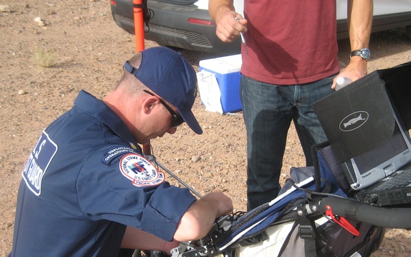 A U.S. Coast Guard member in navy blue uniform is kneeling and working on a wheeled ground-penetrating radar (GPR) cart equipped with a ruggedized Panasonic Toughbook laptop. A second person in civilian clothing (red shirt, jeans) stands nearby observing. The setting appears to be an arid, desert-like outdoor environment with gravel ground and a vehicle visible in the background.