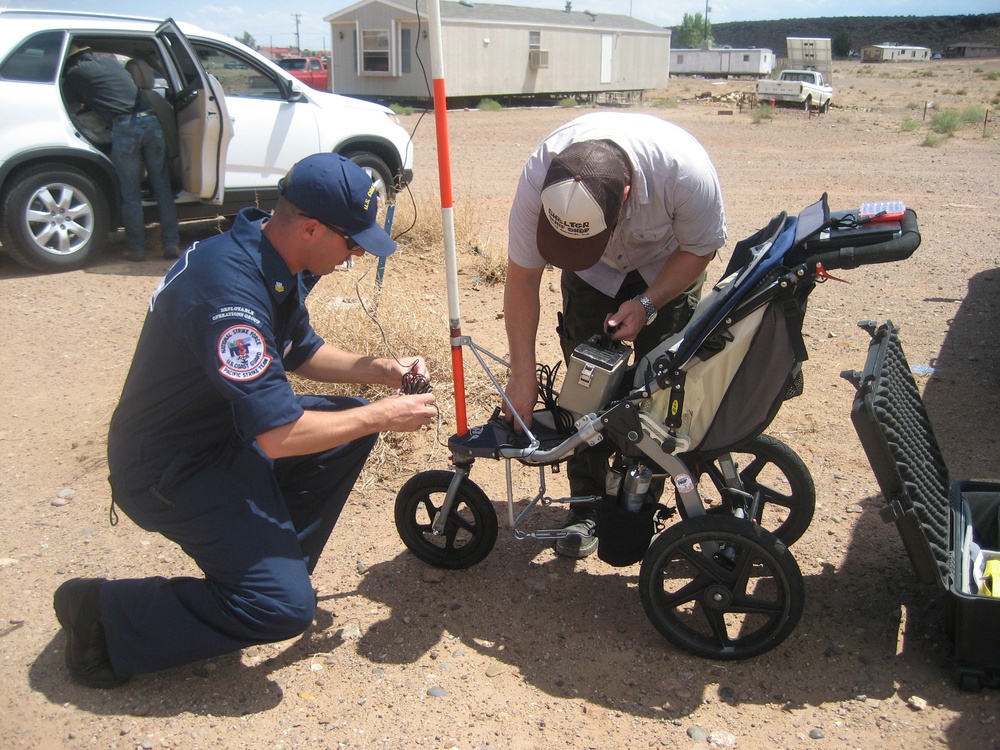 Two individuals are working together to set up or configure a wheeled ground-penetrating radar (GPR) or similar survey equipment in an arid, desert-like environment. One person is wearing a U.S. Coast Guard National Strike Force uniform and cap, while the other wears civilian field attire with a hat. They appear to be at a rural location with mobile homes and vehicles visible in the background.