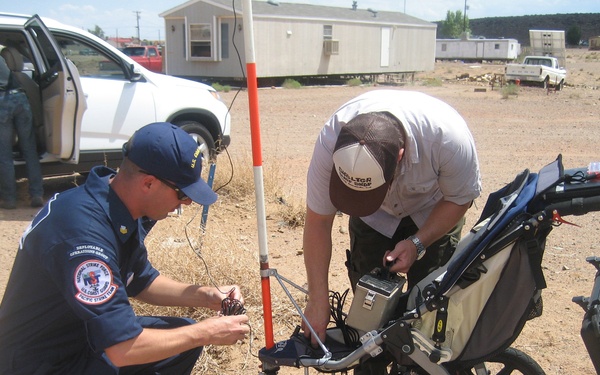 Two individuals are working together to set up or configure a wheeled ground-penetrating radar (GPR) or similar survey equipment in an arid, desert-like environment. One person is wearing a U.S. Coast Guard National Strike Force uniform and cap, while the other wears civilian field attire with a hat. They appear to be at a rural location with mobile homes and vehicles visible in the background.