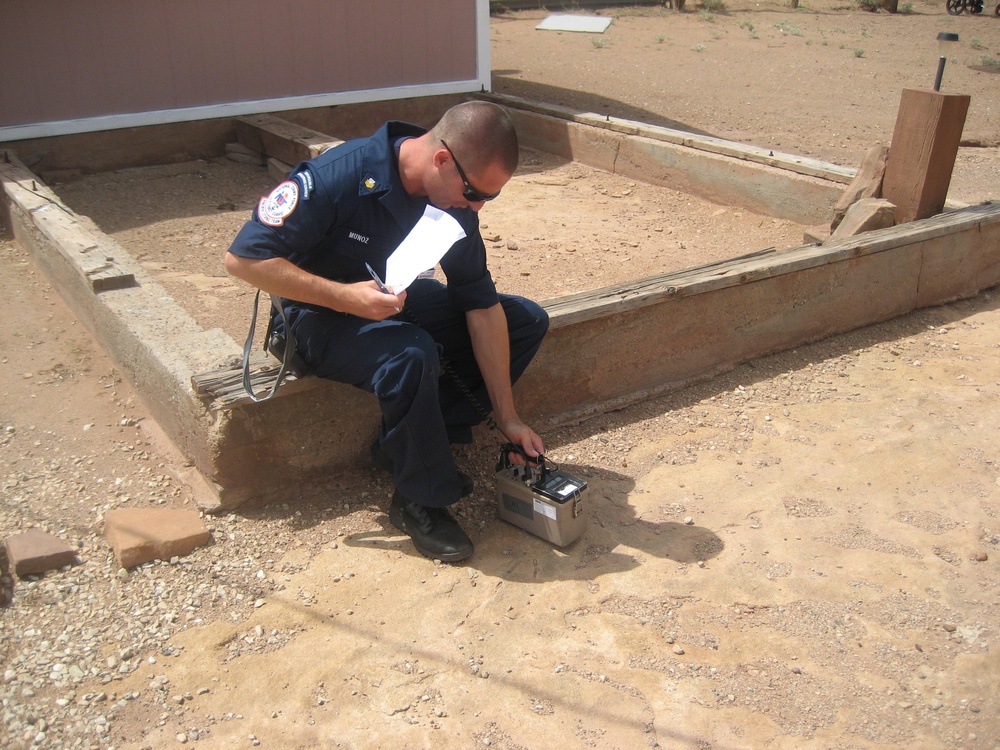 A uniformed officer or firefighter identified as 'MUNOZ' on his name tag is crouching near a concrete foundation or border in an arid, desert-like outdoor environment. He is operating or reading a portable electronic instrument/meter placed on the sandy ground while holding papers in his other hand. The setting appears to be an inspection or assessment of a structure's foundation or site.