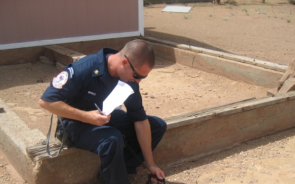 A uniformed officer or firefighter identified as 'MUNOZ' on his name tag is crouching near a concrete foundation or border in an arid, desert-like outdoor environment. He is operating or reading a portable electronic instrument/meter placed on the sandy ground while holding papers in his other hand. The setting appears to be an inspection or assessment of a structure's foundation or site.