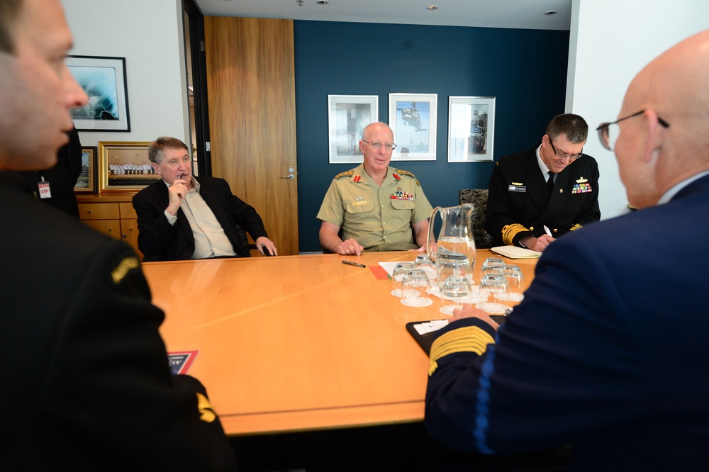 A formal military meeting is taking place around a conference table in a modern office setting. Multiple individuals in military uniforms of different branches are present alongside at least one civilian. The setting appears to be a high-level briefing or diplomatic meeting.