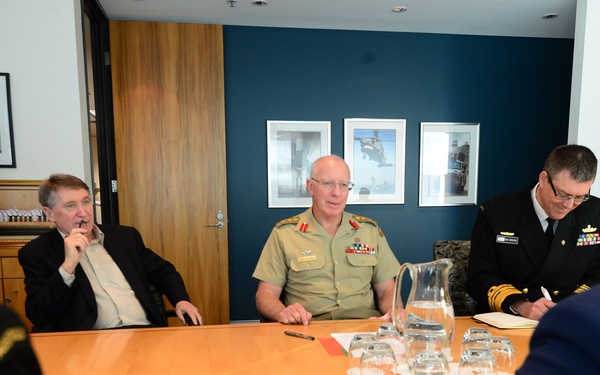 A formal military meeting is taking place around a conference table in a modern office setting. Multiple individuals in military uniforms of different branches are present alongside at least one civilian. The setting appears to be a high-level briefing or diplomatic meeting.