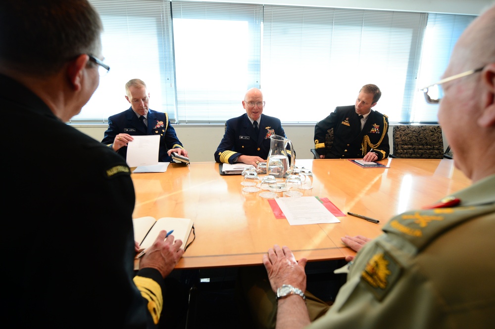 A formal military meeting is taking place around a large wooden conference table. Several uniformed officers from what appear to be different branches and nationalities are seated and engaged in discussion, with one central figure appearing to be speaking. The setting is a professional conference room with large windows in the background.
