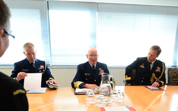 A formal military meeting is taking place around a large wooden conference table. Several uniformed officers from what appear to be different branches and nationalities are seated and engaged in discussion, with one central figure appearing to be speaking. The setting is a professional conference room with large windows in the background.
