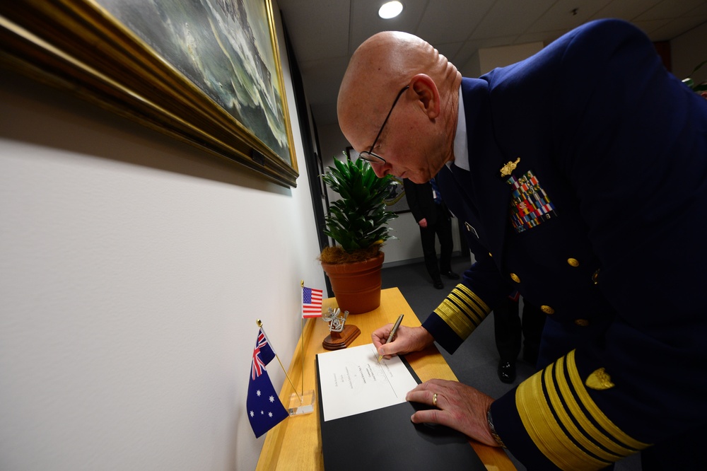 A senior military officer in a dark blue dress uniform with gold sleeve stripes and multiple service ribbons is signing a document at a wooden table. Two small flags — one Australian and one American — are displayed on the table, suggesting a bilateral or diplomatic meeting. A framed maritime painting and a potted plant are visible in the background.