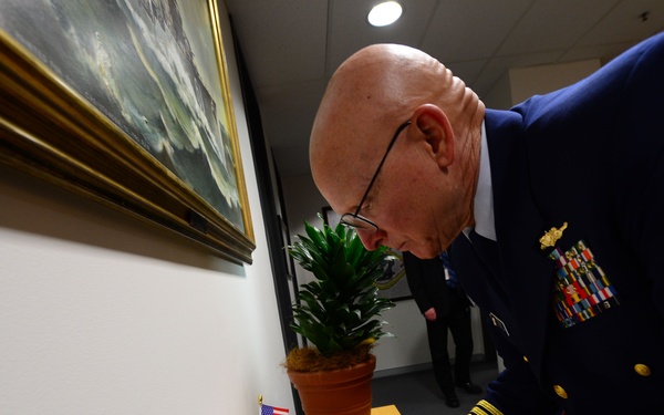 A senior military officer in a dark blue dress uniform with gold sleeve stripes and multiple service ribbons is signing a document at a wooden table. Two small flags — one Australian and one American — are displayed on the table, suggesting a bilateral or diplomatic meeting. A framed maritime painting and a potted plant are visible in the background.