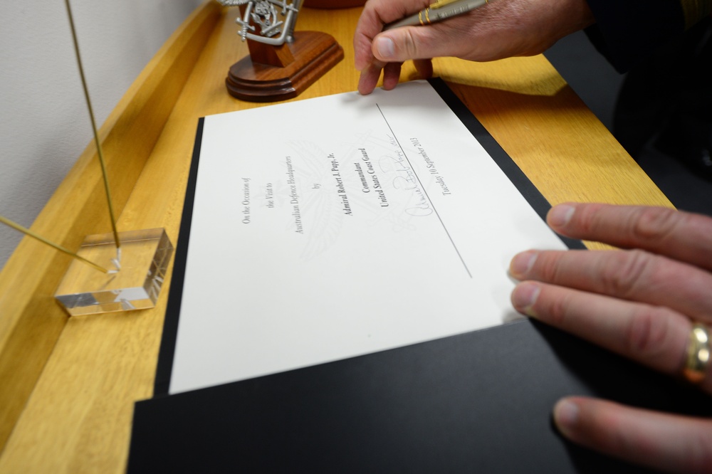 A person is handling a formal document or certificate placed in a black presentation folder on a wooden desk. The document references a visit to Australian Defence Headquarters by Admiral Robert J. Papp, Jr., Commandant of the United States Coast Guard, dated Tuesday 10 September 2013. A signature is visible on the document along with a decorative award or trophy in the background.