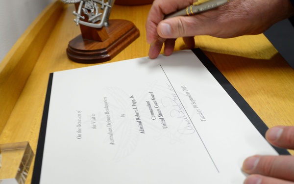 A person is handling a formal document or certificate placed in a black presentation folder on a wooden desk. The document references a visit to Australian Defence Headquarters by Admiral Robert J. Papp, Jr., Commandant of the United States Coast Guard, dated Tuesday 10 September 2013. A signature is visible on the document along with a decorative award or trophy in the background.