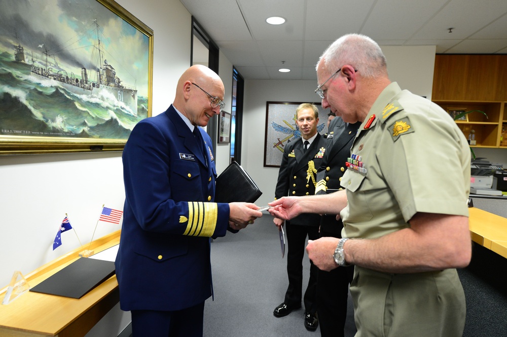 A formal military exchange is taking place in an office setting between personnel from what appears to be the United States Coast Guard and the Australian Defence Force. Two officers in the foreground are exchanging what appears to be a business card or document, while a third officer in dark naval uniform observes in the background. Small Australian and American flags on the desk suggest this is an official bilateral meeting or visit.