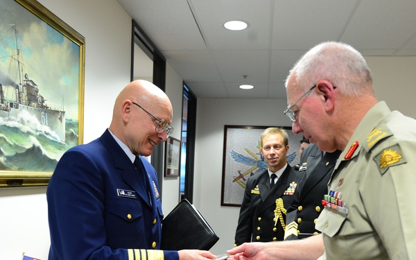 A formal military exchange is taking place in an office setting between personnel from what appears to be the United States Coast Guard and the Australian Defence Force. Two officers in the foreground are exchanging what appears to be a business card or document, while a third officer in dark naval uniform observes in the background. Small Australian and American flags on the desk suggest this is an official bilateral meeting or visit.
