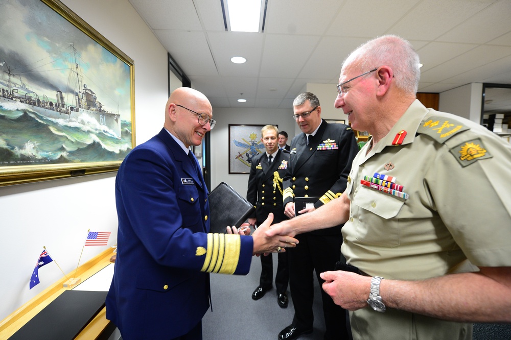 A formal military meeting is taking place indoors, likely in an official office or conference room. A U.S. Navy admiral in dark blue dress uniform is shaking hands with an Australian Army general in tan/khaki service dress uniform. Two additional naval officers in dark uniforms observe the exchange in the background.