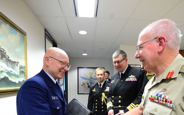 A formal military meeting is taking place indoors, likely in an official office or conference room. A U.S. Navy admiral in dark blue dress uniform is shaking hands with an Australian Army general in tan/khaki service dress uniform. Two additional naval officers in dark uniforms observe the exchange in the background.
