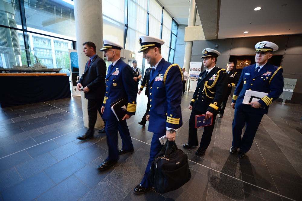 A group of military officers in formal dress uniforms are walking through what appears to be a modern building lobby or conference center. The officers are wearing multiple different uniforms suggesting representation from different branches or nations. The setting appears to be an official military event or conference.