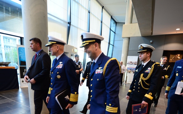A group of military officers in formal dress uniforms are walking through what appears to be a modern building lobby or conference center. The officers are wearing multiple different uniforms suggesting representation from different branches or nations. The setting appears to be an official military event or conference.