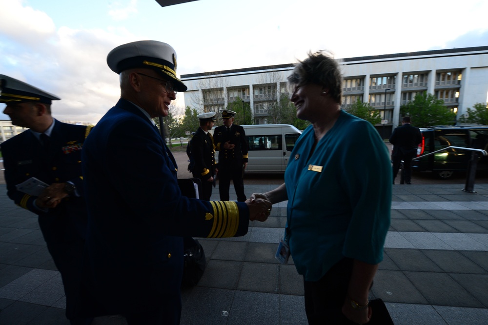 A senior military officer in a dark blue dress uniform with gold sleeve stripes and a white peaked cap is shaking hands with a civilian woman wearing a teal blouse and a name badge. The setting appears to be an outdoor plaza or entrance area of an institutional building, likely during an official reception or arrival ceremony. Several other uniformed personnel are visible in the background near parked vehicles.