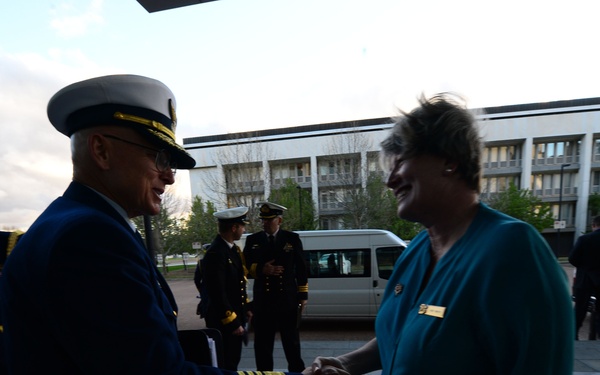 A senior military officer in a dark blue dress uniform with gold sleeve stripes and a white peaked cap is shaking hands with a civilian woman wearing a teal blouse and a name badge. The setting appears to be an outdoor plaza or entrance area of an institutional building, likely during an official reception or arrival ceremony. Several other uniformed personnel are visible in the background near parked vehicles.