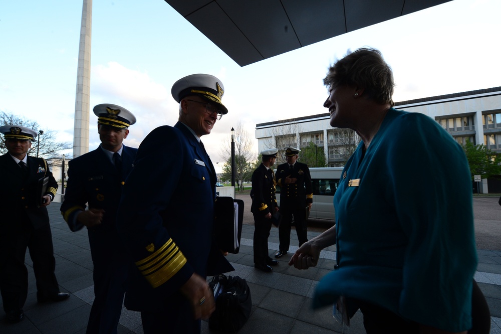 A group of military officers in dress uniforms are gathered outside a government or institutional building. A senior officer with gold sleeve stripes and a white-topped cap is engaged in a friendly conversation with a civilian woman in a teal jacket. The setting appears to be an official visit or formal reception.