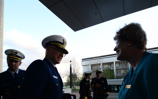A group of military officers in dress uniforms are gathered outside a government or institutional building. A senior officer with gold sleeve stripes and a white-topped cap is engaged in a friendly conversation with a civilian woman in a teal jacket. The setting appears to be an official visit or formal reception.