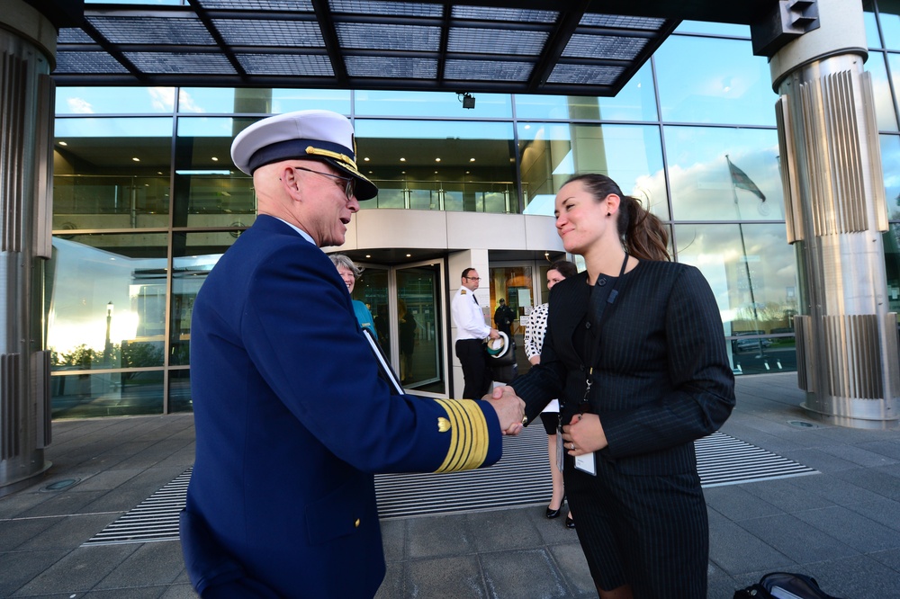 A senior military officer in a dark blue dress uniform with gold sleeve stripes and a white peaked cap is shaking hands with a woman in a black pinstripe business suit outside a modern glass-fronted building. The formal greeting appears to be an official reception or welcoming ceremony. Several other individuals, including another uniformed officer, are visible in the background near the building entrance.