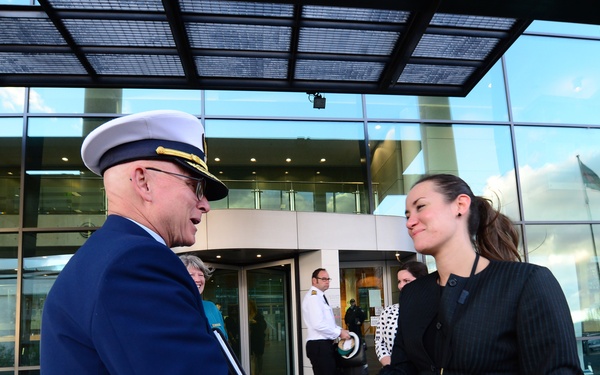 A senior military officer in a dark blue dress uniform with gold sleeve stripes and a white peaked cap is shaking hands with a woman in a black pinstripe business suit outside a modern glass-fronted building. The formal greeting appears to be an official reception or welcoming ceremony. Several other individuals, including another uniformed officer, are visible in the background near the building entrance.