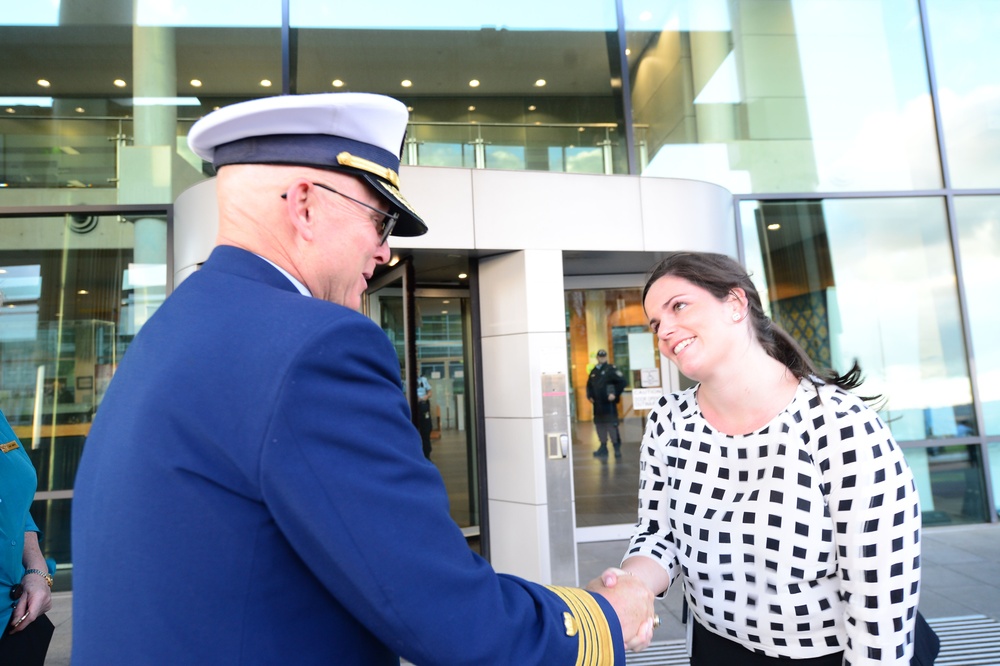 A uniformed military or coast guard officer is shaking hands with a woman in a black-and-white checkered top outside a modern glass building. The meeting appears formal and cordial, likely an official greeting or diplomatic encounter. The setting suggests a government or institutional facility.