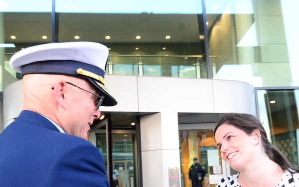A uniformed military or coast guard officer is shaking hands with a woman in a black-and-white checkered top outside a modern glass building. The meeting appears formal and cordial, likely an official greeting or diplomatic encounter. The setting suggests a government or institutional facility.