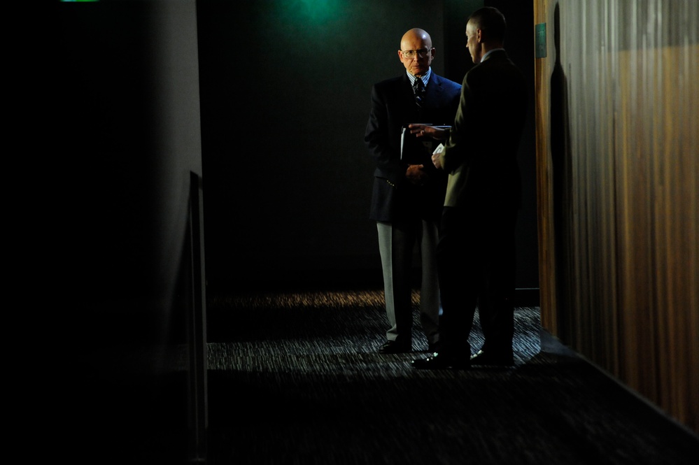 Two men in formal business attire are conversing in a dimly lit corridor or hallway. One man, who is bald and wearing glasses, is facing the camera while holding documents or a folder. The scene has dramatic, high-contrast lighting with a green ambient light visible in the upper background.