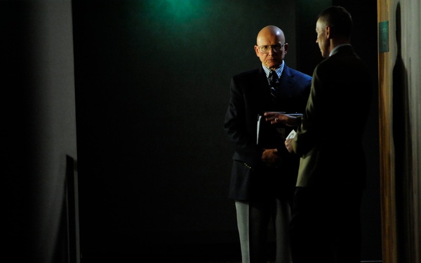 Two men in formal business attire are conversing in a dimly lit corridor or hallway. One man, who is bald and wearing glasses, is facing the camera while holding documents or a folder. The scene has dramatic, high-contrast lighting with a green ambient light visible in the upper background.