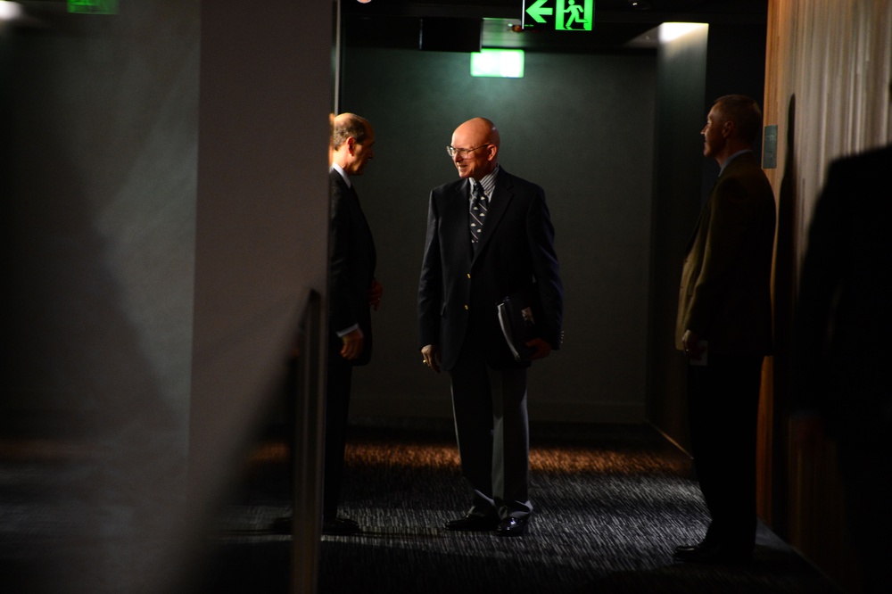 Three men in formal business attire are gathered in what appears to be a dimly lit corridor or hallway near an emergency exit. The central figure is a bald man wearing glasses and a dark suit, holding documents or a folder, appearing to be engaged in conversation. The scene suggests a professional or official setting, possibly before or after a formal meeting.
