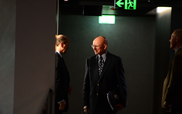 Three men in formal business attire are gathered in what appears to be a dimly lit corridor or hallway near an emergency exit. The central figure is a bald man wearing glasses and a dark suit, holding documents or a folder, appearing to be engaged in conversation. The scene suggests a professional or official setting, possibly before or after a formal meeting.