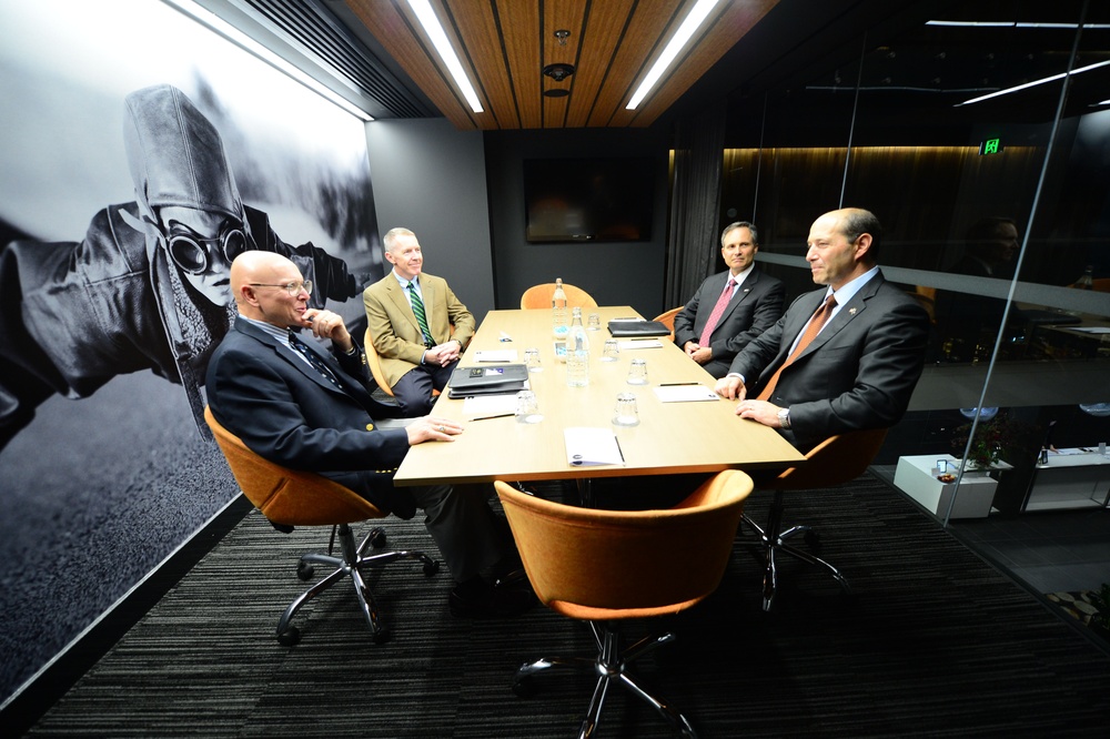 Four men in business attire are seated around a rectangular conference table in a modern, upscale meeting room. The room features a large black-and-white mural of a football player on one wall, warm wood ceiling panels, and glass partition walls. The men appear to be engaged in a formal discussion or meeting.