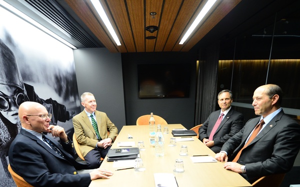 Four men in business attire are seated around a rectangular conference table in a modern, upscale meeting room. The room features a large black-and-white mural of a football player on one wall, warm wood ceiling panels, and glass partition walls. The men appear to be engaged in a formal discussion or meeting.