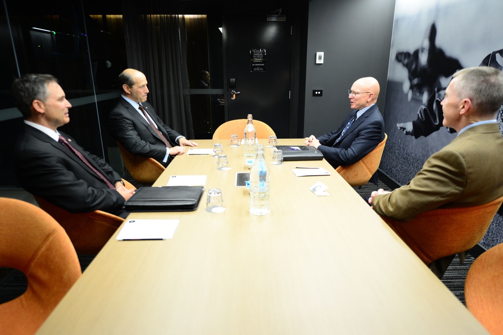 Four men in formal business attire are seated around a rectangular conference table in a modern meeting room, engaged in what appears to be a formal bilateral or diplomatic discussion. The room features dark walls with what appears to be a military aircraft mural on the right side, orange upholstered chairs, and a sign on the door indicating room capacity. Documents, water bottles, glasses, and a laptop are visible on the table.